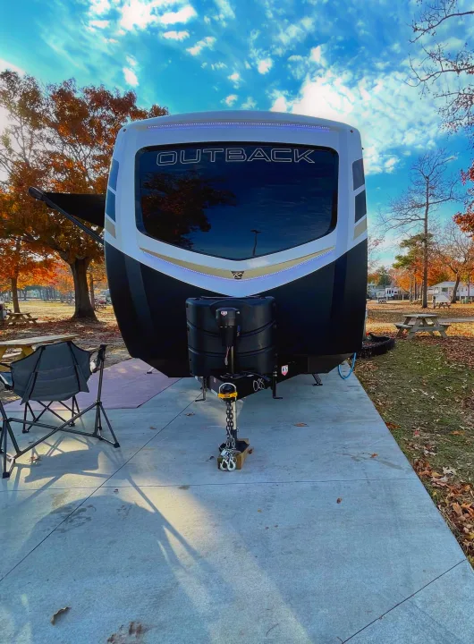 Front view of an 'Outback' travel trailer RV rental at a sunny autumn campground, with chairs and fall foliage.. Keystone RV Outback 2021