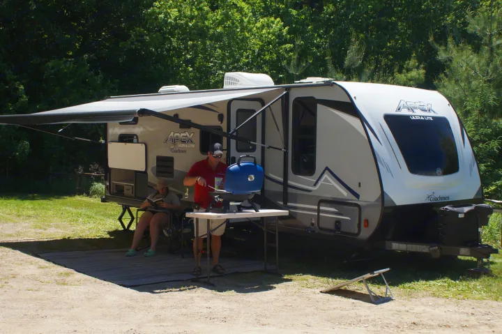 Relaxing RV rental: A man grills and a woman reads under the awning of their Coachmen Apex travel trailer at a campsite.. Apex Nano 245 BHS 2020
