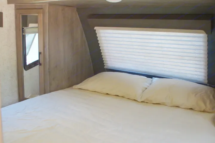 Inside an RV rental, a compact bedroom features a bed with white sheets, two pillows, and a window with horizontal blinds.. Apex Nano 245 BHS 2020