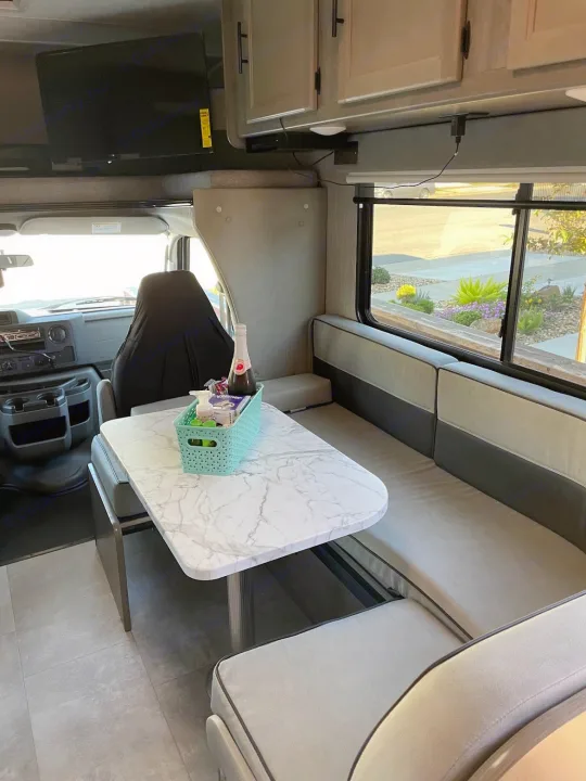 Inside a modern RV rental: a bright dining area with table, bench seats, overhead TV, and cabinets.. Forest River Coachmen Freelander 2021