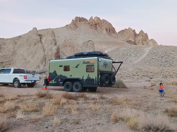 Rugged RV rental hitched to a white truck in a vast desert landscape with rocky mountains. A child explores nearby.. Xplorer X22 2022