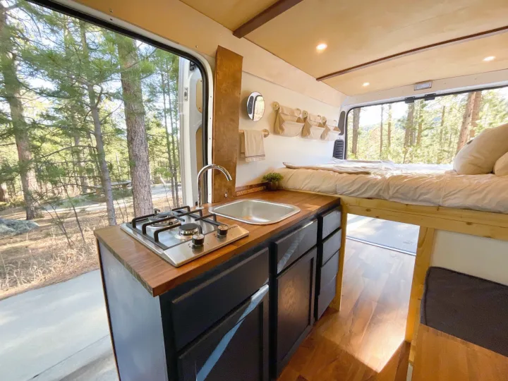 Bright rv rental camper van kitchen & bed with forest view. Features stove, sink, & wood accents for cozy travel.. Other Dodge 2021