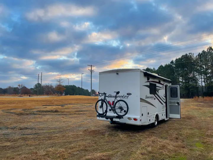 A white Sunseeker RV rental, with a bicycle on its rear rack, parked in an open field under a cloudy sky.. Forest River Sunseeker 2017