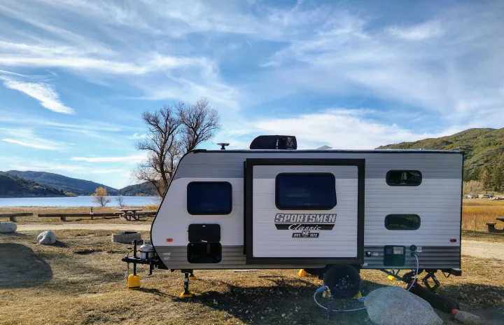 Scenic RV rental setup: a Sportsmen Classic travel trailer by a lake, surrounded by autumn trees and mountains, perfect for camping.. KZ 181BH 2021