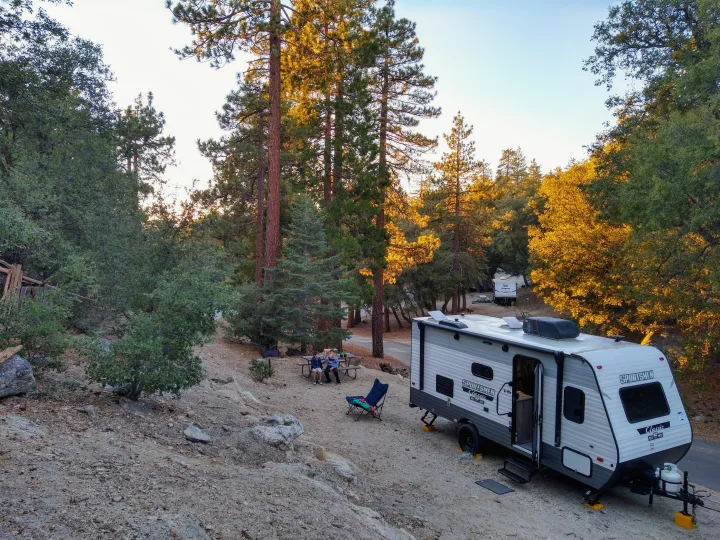 Campsite fun with an RV rental: A travel trailer parked amidst trees, with two people at a picnic table.. KZ 181BH 2021