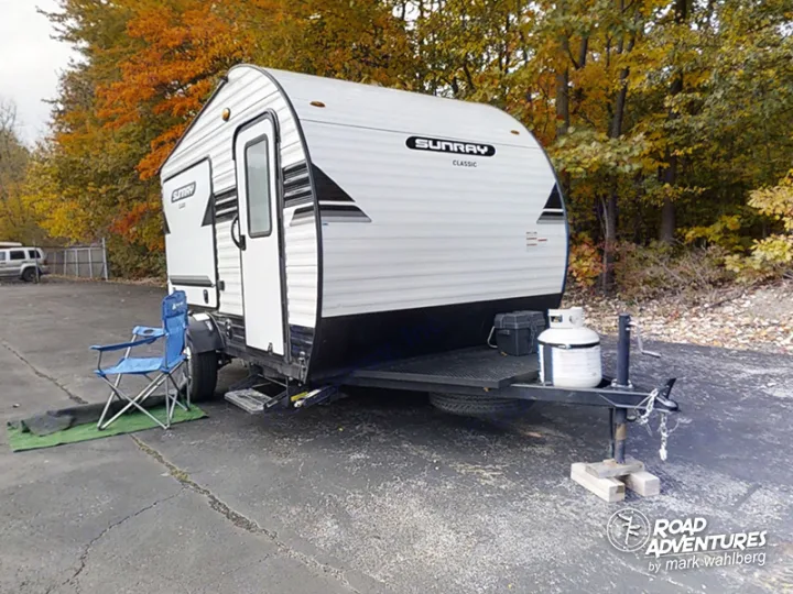 This white Sunray Classic travel trailer, an rv rental, is set up for autumn road trip adventures with a chair.. Sunset Park Sunray 2021