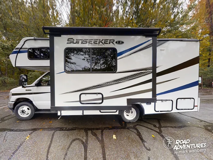 Side view of a white Forest River Sunseeker rv rental with blue/gray decals & extended awning, parked among trees.. Forest River Sunseeker 2022