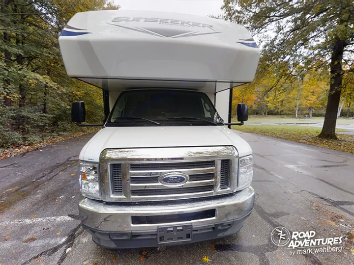 Front view of a white Ford Sunseeker RV rental parked on an asphalt road, surrounded by trees with autumn foliage.. Forest River Sunseeker 2022