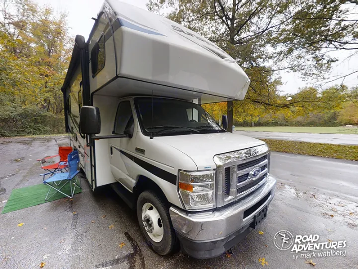 White RV rental parked on a paved lot at a fall campsite. Red & blue chairs with green mat outside for cozy road adventures.. Forest River Sunseeker 2022