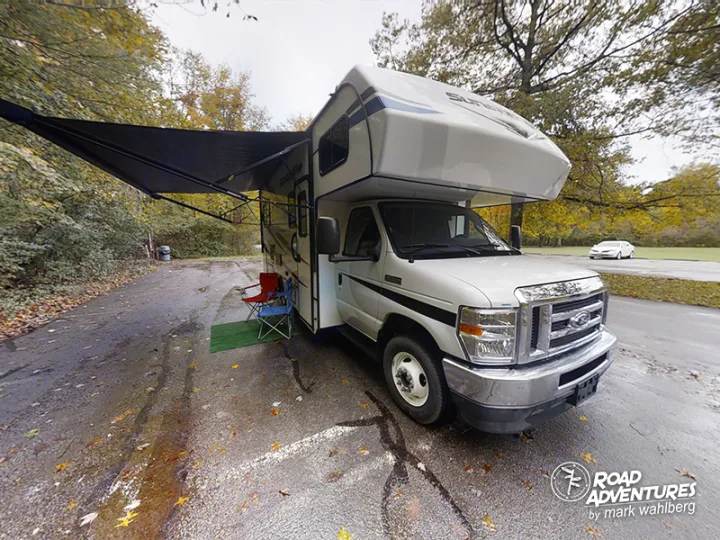 Spacious white rv rental parked with awning and chairs set up on an asphalt lot, surrounded by trees. Ideal for camping trips.. Forest River Sunseeker 2022