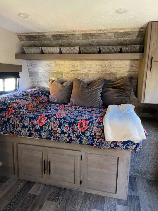 Cozy RV rental bedroom featuring a bed with a floral quilt, pillows, overhead shelves, and under-bed storage.. Keystone RV Springdale 2021