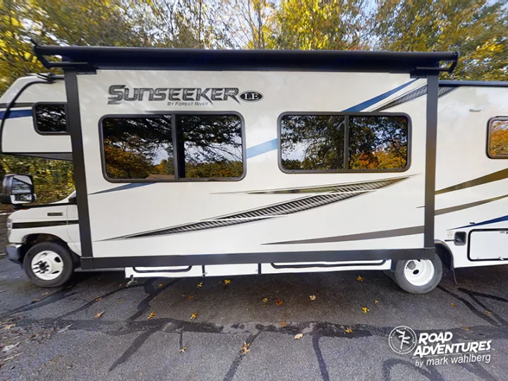 Side view of a white and blue Sunseeker RV rental motorhome with awning extended, parked on asphalt near trees.. Forest River Sunseeker 2022