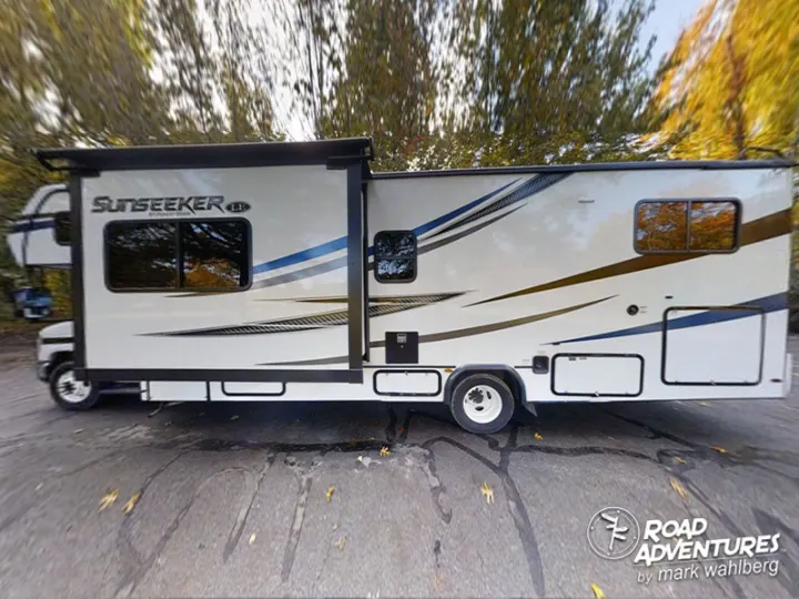 Side view of a Sunseeker rv rental, a white Class C motorhome with blue/gold accents, parked by trees.. Forest River Sunseeker 2022