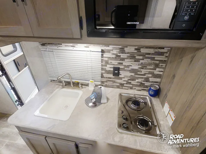 Compact RV rental kitchen with sink, two-burner stove, microwave, and cabinets. Ideal for travel cooking needs.. Gulf Stream Amerilite 2021