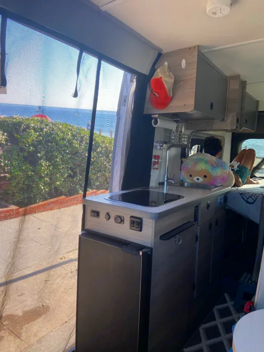 This RV rental offers ocean views! A person relaxes inside a cozy camper van with a compact kitchen and sink setup.. Winnebago Solis 2021