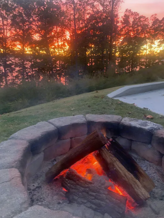 Cozy RV rental evening: a crackling fire in a stone pit illuminates logs against a vibrant sunset.. Keystone RV Springdale 2021