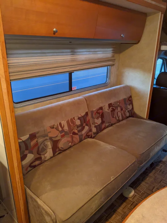 RV rental living space: a long tan sofa with patterned back cushions, wood cabinets, and a window showing an EXIT sign.. Winnebago Itasca Navion 2008