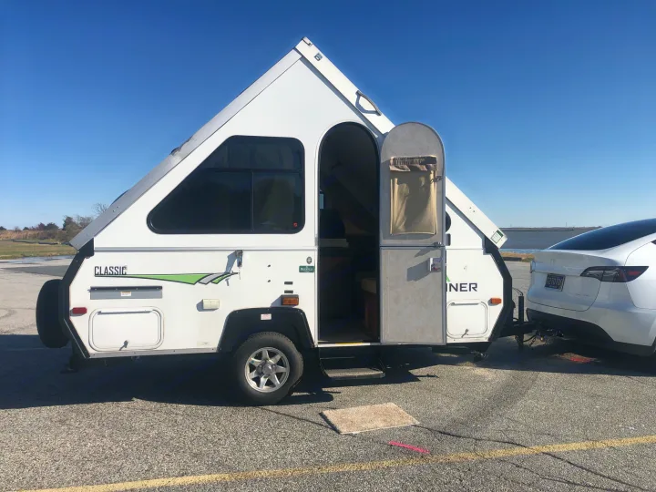 A white A-frame **rv rental** camper is hitched to a Tesla, parked outdoors with its door open.. A Liner A Liner Popup Trailer 2014