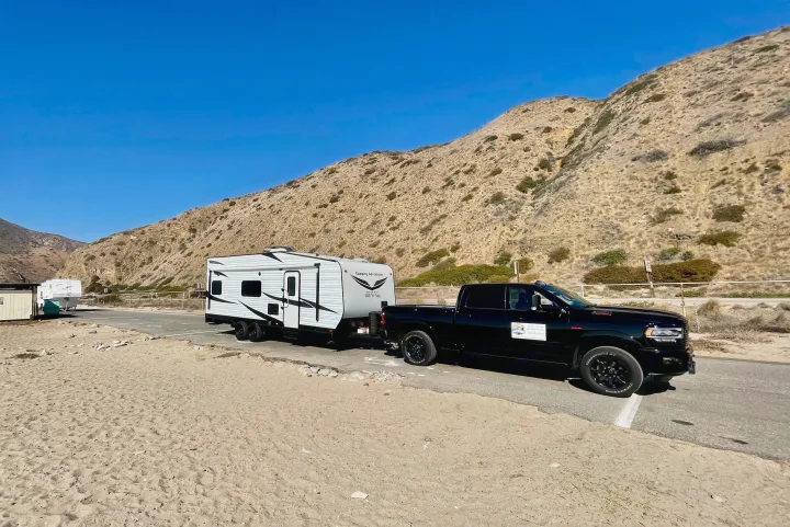 An RV rental, a white travel trailer pulled by a black truck, parked by sand & dry hills under clear blue sky.. Dune Sport Toy Hauler 2017