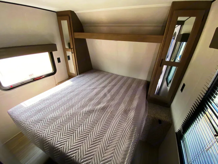 A well-appointed RV rental bedroom features a textured bedspread, built-in storage cabinets, and bright windows.. Keystone Hideout 2021