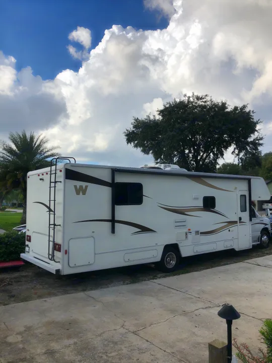 White RV rental, a recreational vehicle with a rear ladder & awning, parked on a driveway under a partly cloudy sky.. Winnebago Minnie Winnie 2014