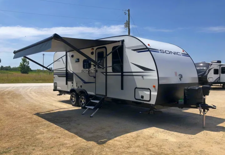 Modern white Sonic travel trailer with extended awning, ready for rv rental. Parked on a gravel lot under a clear sky.. Sonic Sonic Trailer 2025
