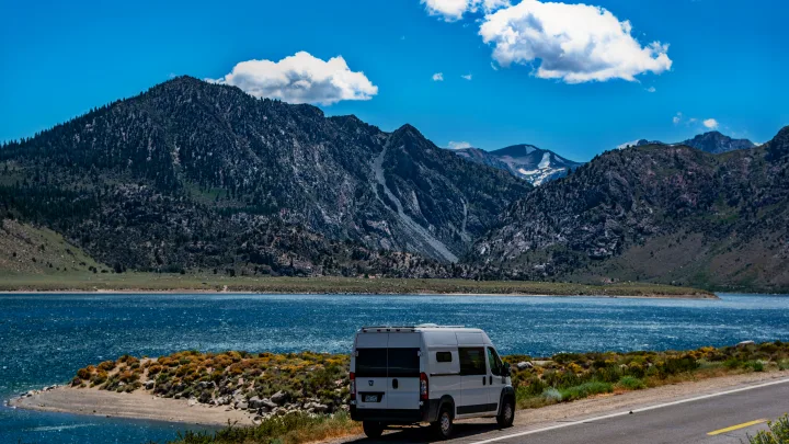 A white RV rental parked on a scenic road beside a shimmering blue lake, with towering mountains and a clear sky.. RAM Promaster 1500 2017