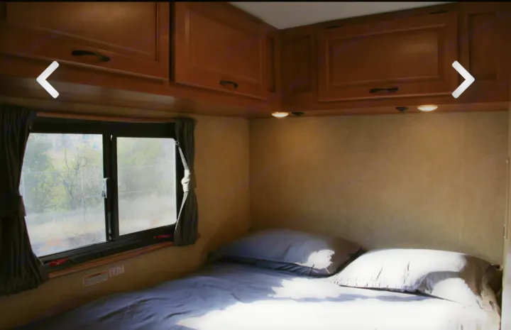 Cozy rv rental bedroom featuring a made bed, large window, and wooden overhead storage cabinets.. Thor Majestic 2017