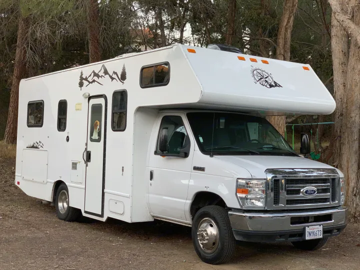 White Ford RV rental parked among trees, perfect for camping. A cute beagle peeks out from the entrance door.. Thor Majestic 2017