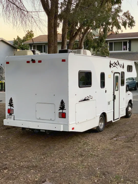 White RV rental with mountain decals, parked on dirt. A dog peeks from the door, ready for camping adventures.. Thor Majestic 2017