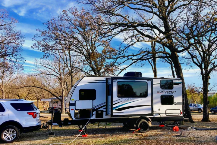 White SUV towing a Forest River Surveyor Legend rv rental at a fall campsite with large, bare trees under a blue sky.. Forest River Surveyor Legend 2022