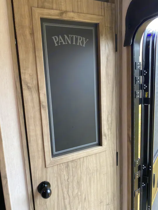 RV rental interior: A light wood-grain pantry door with 'Pantry' signage stands next to the open RV entrance.. Forest River Cherokee Alpha Wolf 2021