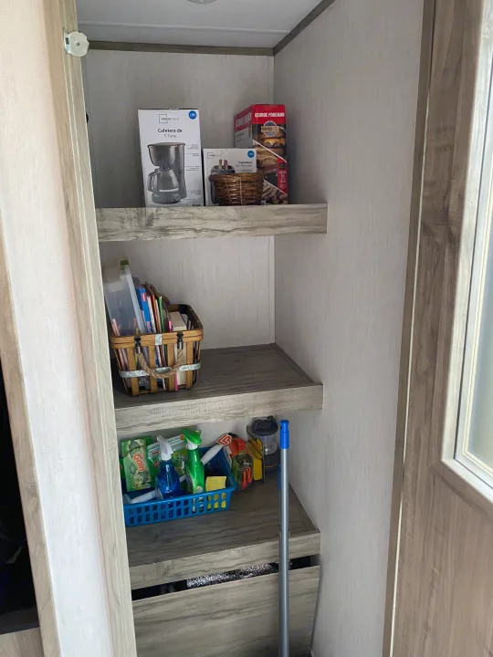 RV rental storage: Shelves stocked with a coffee maker, grill, books, and cleaning supplies; a mop leans nearby.. Forest River Cherokee Alpha Wolf 2021