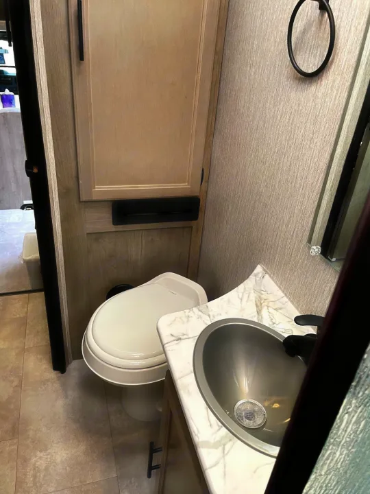 Compact RV rental bathroom featuring a white toilet, gray sink, and wood-toned cabinet. Essential travel amenity.. Forest River Coachmen Freelander 2021