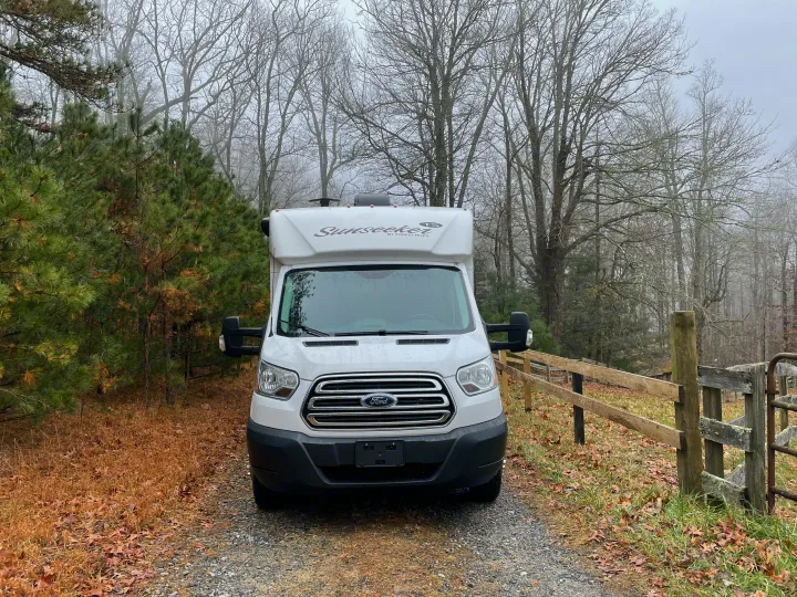 White Ford Sunseeker RV rental on a gravel driveway, flanked by pines and a wooden fence, with foggy bare trees behind.. Forest River Sunseeker 2017