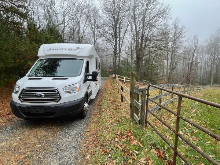 White Sunseeker RV rental parked on a gravel drive, with a rustic fence and bare trees in the misty, overcast woods.. Forest River Sunseeker 2017