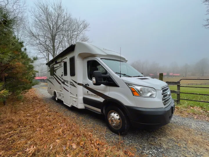 White motorhome RV rental on a damp gravel path amidst bare trees & misty fields, suggesting a serene outdoor escape.. Forest River Sunseeker 2017