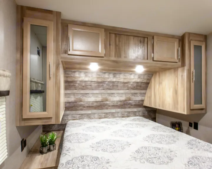 Cozy RV rental bedroom with light-patterned bed, rustic wood-look headboard, and overhead cabinets. Ideal for travel.. Forest River Coachmen Catalina Legacy 2021