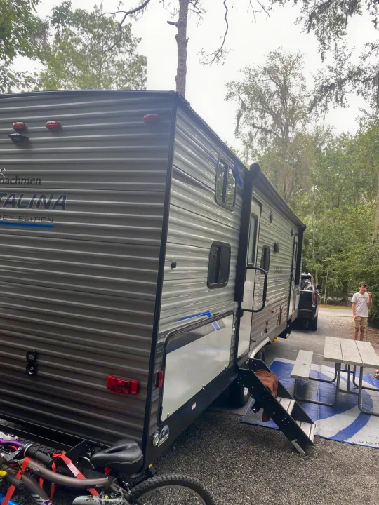 This RV rental, a gray Coachmen Catalina travel trailer, is at a shaded campsite with a picnic area, steps, and bicycles.. Forest River Coachmen Catalina Legacy 2021