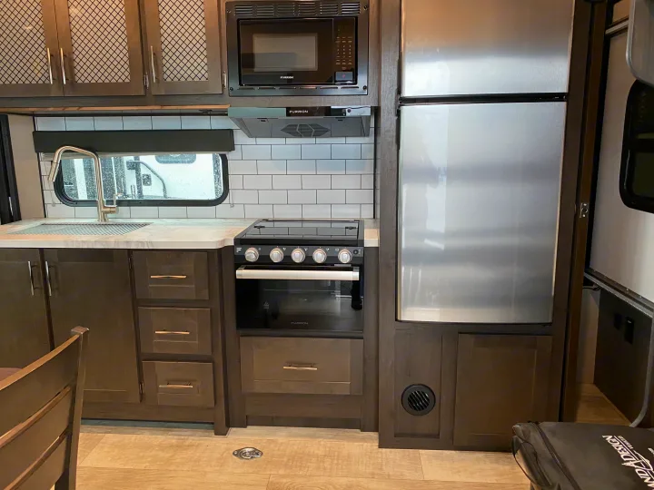 Modern RV rental kitchen with stainless steel appliances like a microwave, stove, fridge, and sink. Dark wood cabinets.. Grand Design Momentum 2021