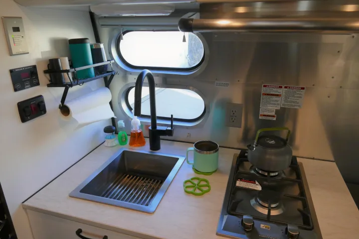 Compact kitchen in an RV rental, featuring a sink, two-burner stove, and monitoring displays for battery/water tanks.. Airstream Caravel 2020
