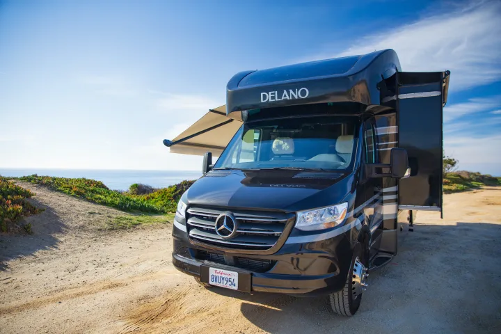 Black Mercedes Sprinter camper van for RV rental, with awning and slide-out extended, parked on a dirt path by the ocean.. Thor Delano Sprinter 2020