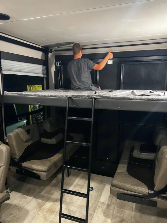 Inside an RV rental, a man sits on a bunk bed, overlooking convertible seating in a toy hauler garage. Ladder provides access.. Grand Design Momentum 2021