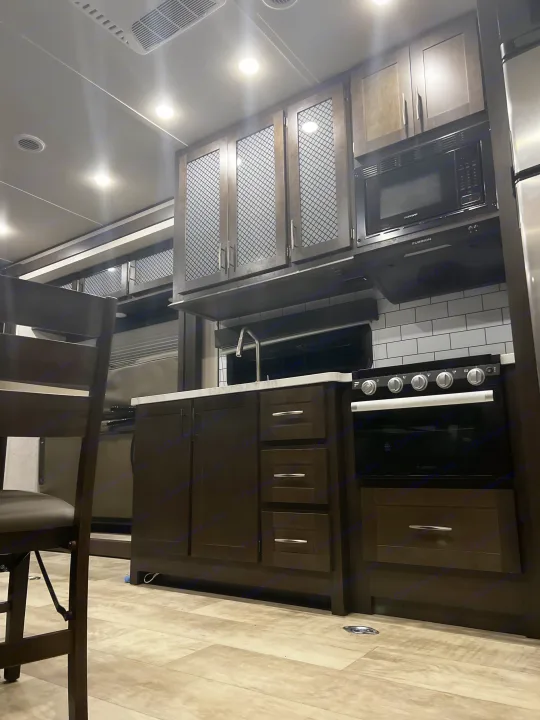 A modern RV rental kitchen showcases dark cabinetry, white countertops, an oven, microwave, and ample overhead storage.. Grand Design Momentum 2021