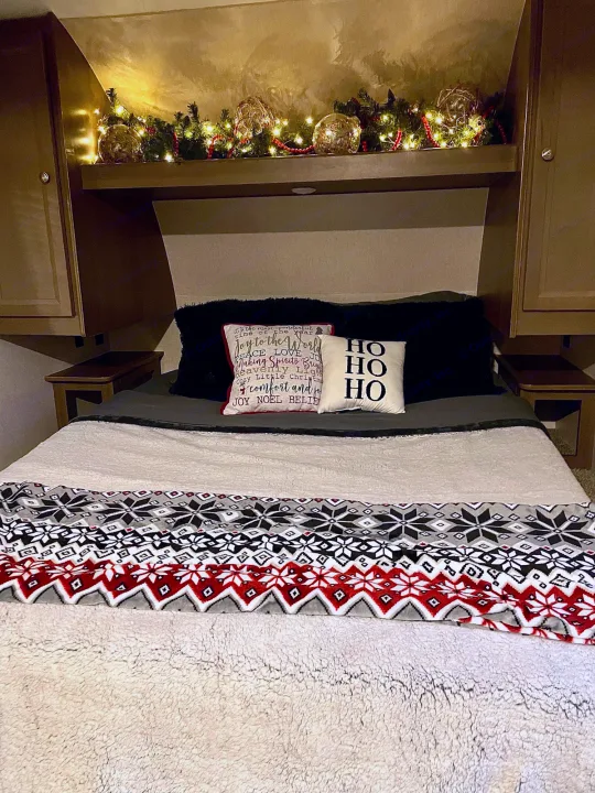 Inside a cozy RV rental: a festive Christmas bedroom with twinkling lights, holiday pillows, and a warm blanket.. Forest River Independence Trail 2021