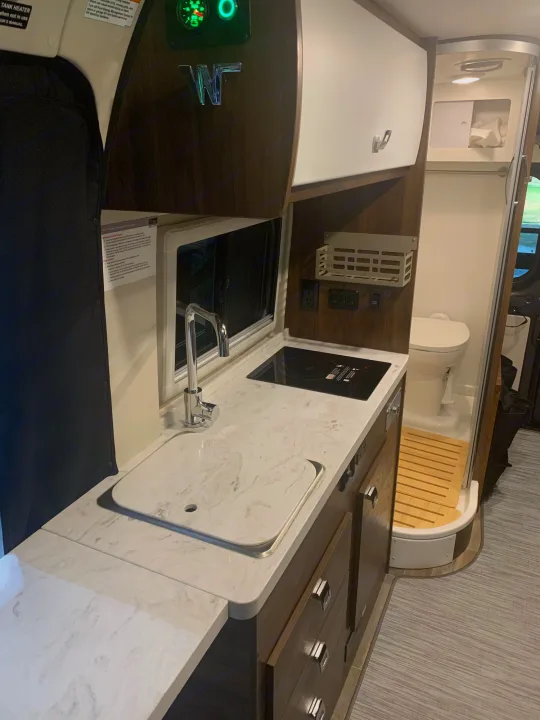 An RV rental's compact interior: modern kitchen with sink & cooktop, plus a wet bath with toilet and wooden mat.. Winnebago Travato 2022