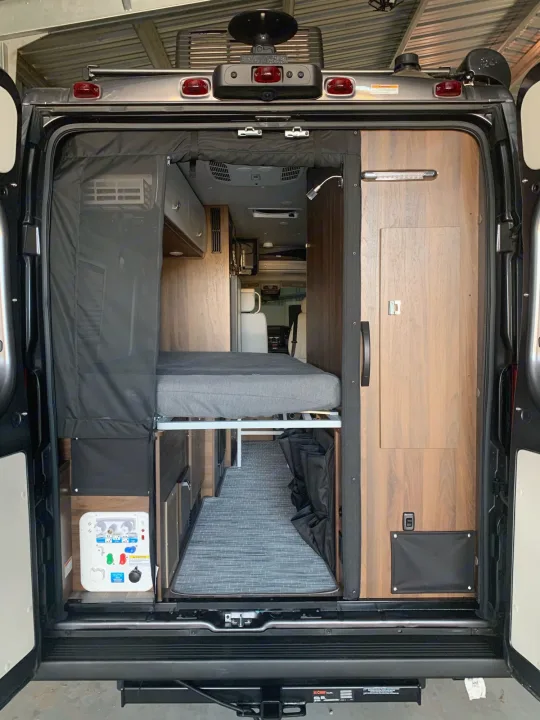 Interior of an RV rental, showing the rear bed, wood cabinetry, and utility panel, viewed from the open back doors.. Winnebago Travato 2022