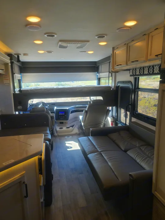 Inside a bright RV rental: view of the front cockpit, comfy seating, overhead bed, and wood floors. Overhead lights are on.. Entegra Vision 29F 2022