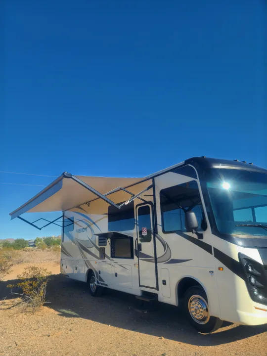 Side view of a white and black RV rental with an extended awning, parked on a dirt lot under a bright blue sky.. Entegra Vision 29F 2022