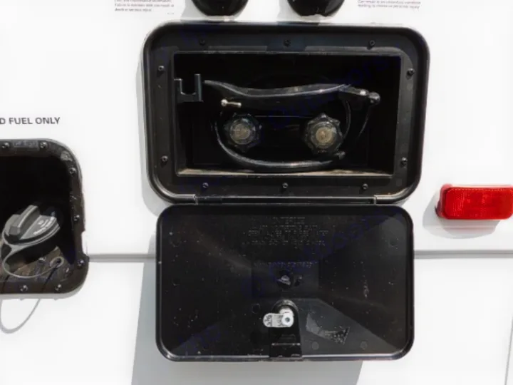 An RV rental's side shows a fuel cap & an open black compartment with an outdoor shower: hose, shower head, and knobs.. Entegra Coach Odyssey 2019